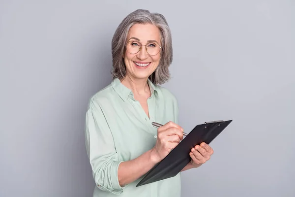 Photo de heureuse souriante belle femme joyeuse dans des lunettes écriture dans le presse-papiers isolé sur fond de couleur grise — Photo