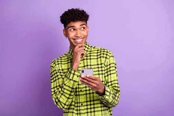 Photo of minded dreamy afro american man hold phone look empty space imagine post isolated on violet color background — Stock fotografie