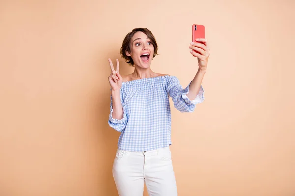 Фотопортрет збудженої дівчини, що показує v-знак тримає телефон в одній руці ізольовано на пастельному бежевому фоні — стокове фото