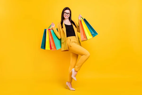 Фото красивої молодої леді одягнені формальні окуляри Блейзер посміхаючись тримає барвисті сумки ізольовані жовтого кольору фону — стокове фото