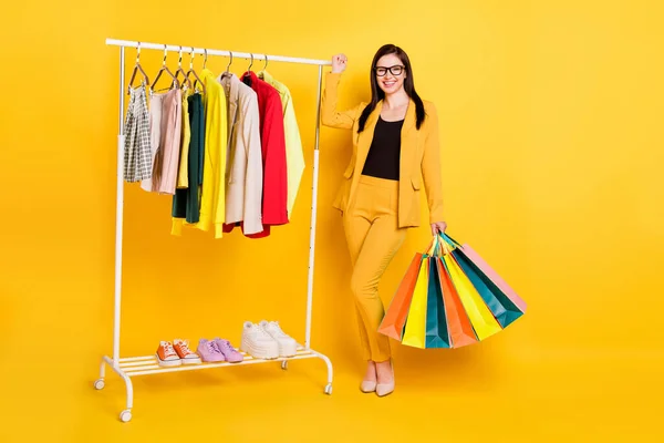 Foto de cuerpo completo de la señora milenaria con bolsos elegir tela en garderobe desgaste gafas stilettos traje aislado sobre fondo amarillo —  Fotos de Stock