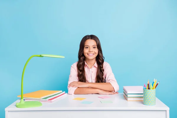 Porträtt av positiv skola flicka sitter bakom skrivbord vikta armar slitage prickade isolerad på blå färg bakgrund — Stockfoto