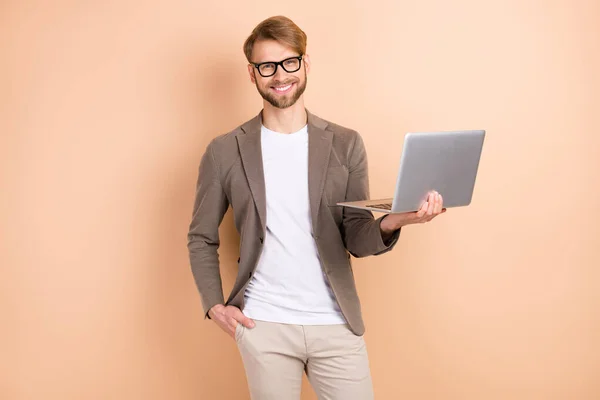 Foto de otimista agradável cabelo loiro homem segurar laptop desgaste óculos casaco cinza isolado no fundo cor bege — Fotografia de Stock