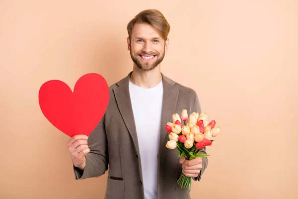 Foto de guapo lindo caballero joven vestido chaqueta celebración tulipanes manojo grande corazón rojo aislado color beige fondo — Foto de Stock