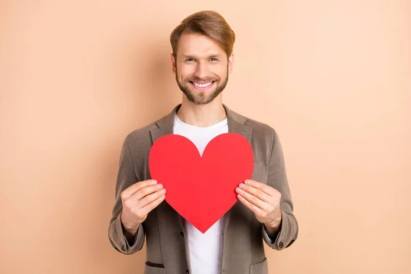 Foto di classe attraente giovane gentiluomo vestito blazer tenendo grande cuore rosso spazio vuoto isolato colore beige sfondo — Foto Stock