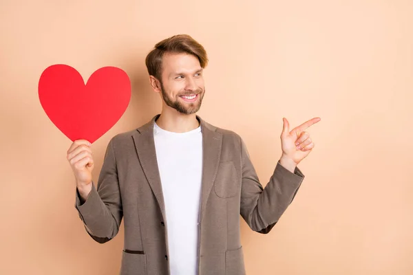 Foto de bonito encantador jovem desgaste casaco sorrindo segurando grande coração vermelho olhando apontando espaço vazio isolado cor bege fundo — Fotografia de Stock