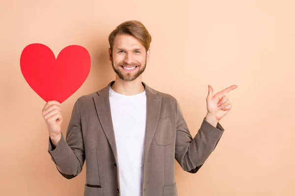 Foto di grazioso signore di classe vestito blazer tenendo grande cuore rosso indicando dito vuoto spazio isolato colore beige sfondo — Foto Stock