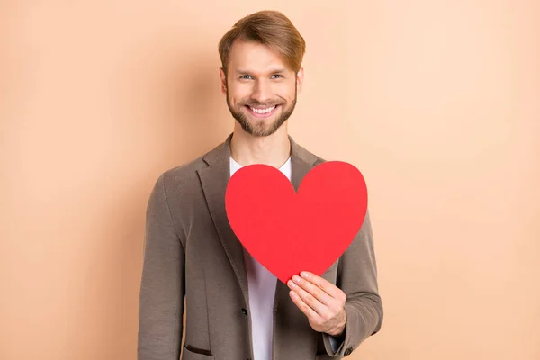 Foto de alegre doce jovem desgaste casaco sorrindo segurando grande coração vermelho isolado cor bege fundo — Fotografia de Stock