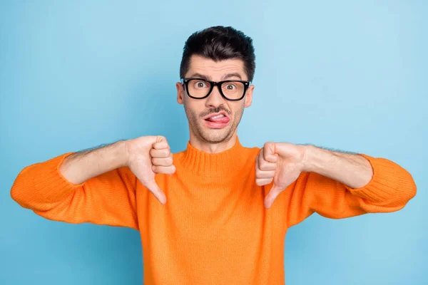 Foto de hombre joven triste triste triste mordida lengua dientes muestran pulgar hacia abajo mala calidad aversión anuncio aislado sobre fondo de color azul —  Fotos de Stock