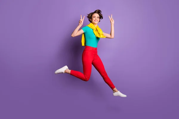 Full längd foto av flicka hoppa visar v-tecken bära bunden tröja t-shirt byxor sneakers isolerad lila färg bakgrund — Stockfoto