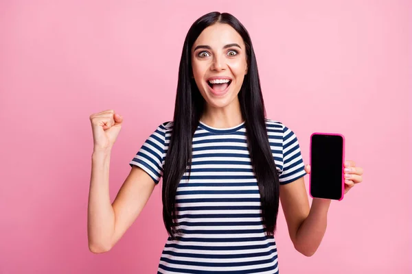 Photo portrait of celebrating girl with raised fist holding phone with blank space in one hand isolated on pastel pink colored background — Stock Photo, Image