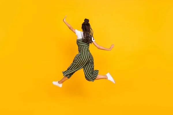 Volledige lengte foto van haasten zorgeloze donkere huid dame rug dragen gestreepte kleding springen hoog geïsoleerde gele kleur achtergrond — Stockfoto