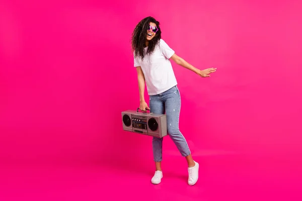 Full size photo of cute lady hold boom box look empty space wear glasses t-shirt jeans sneakers isolated on pink background — стоковое фото