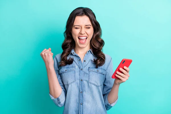 Фото шокованої щасливої молодої жінки в джинсовій сорочці, що читає сучасний пристрій піднімається кулаком ізольований бірюзовий кольоровий фон — стокове фото
