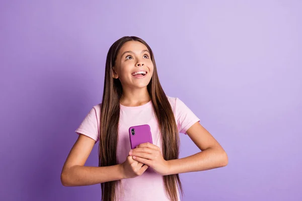 Foto de dulce colegiala impresionada vestida camiseta rosa sosteniendo dispositivo moderno buscando espacio vacío aislado color púrpura fondo — Foto de Stock