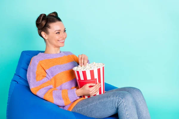 Портрет привлекательной веселой девушки, сидящей в кресле и питающейся кукурузой, смотрящей сериал, развлекаясь на ярко-бирюзовом фоне — стоковое фото