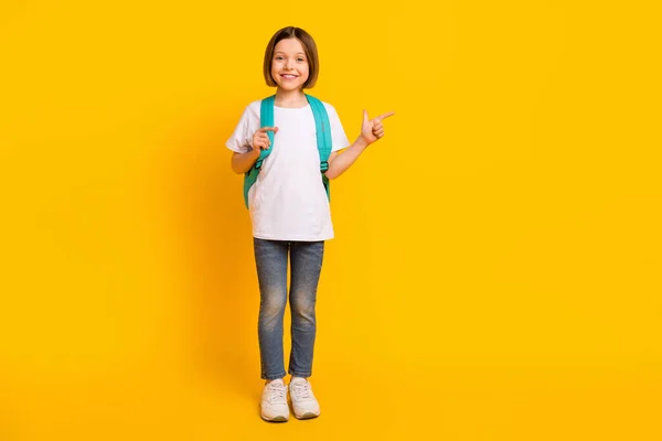Foto di lucido adorabile studentessa vestito bianco vestito zaino indicando dito vuoto spazio sorridente isolato colore giallo sfondo — Foto Stock