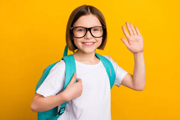 Foto von freundlichen glänzenden Schulmädchen gekleidet weißen Outfit Brille Rucksack winken Arm Handfläche lächelnd isoliert gelbe Farbe Hintergrund — Stockfoto