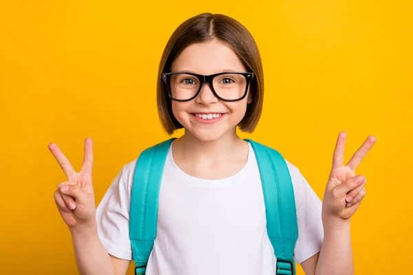 Foto portret klein schoolmeisje glimlachend dragen bril tonen v-teken gebaar geïsoleerde levendige gele kleur achtergrond — Stockfoto