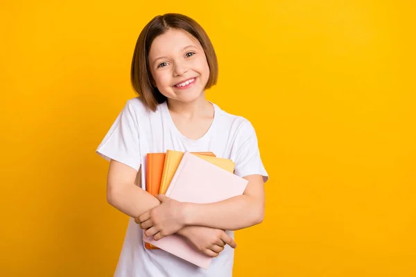 Foto van lieve mooie school meisje dragen witte t-shirt glimlachen knuffelen boek stapel geïsoleerde gele kleur achtergrond — Stockfoto