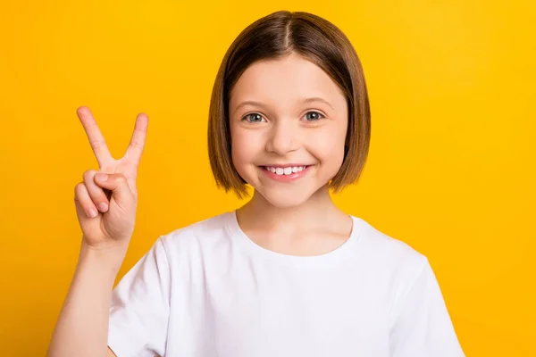 Foto de bonito encantador escola menina desgaste branco t-shirt sorrindo mostrando v-sinal isolado cor amarela fundo — Fotografia de Stock