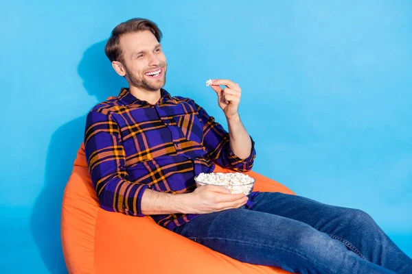 Ritratto di attraente ragazzo allegro seduto sulla sedia mangiare pop corn guardare spettacolo televisivo isolato su vivido sfondo di colore blu — Foto Stock