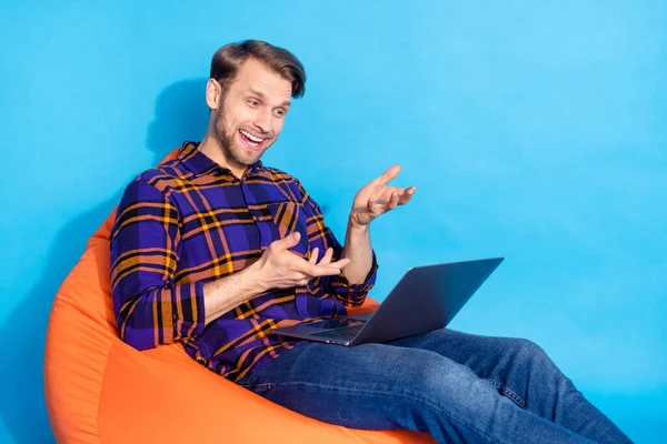 Portrett av en kjekk, munter fyr som sitter i stolen og bruker en bærbar datamaskin som snakker isolert på livaktig blå bakgrunn – stockfoto