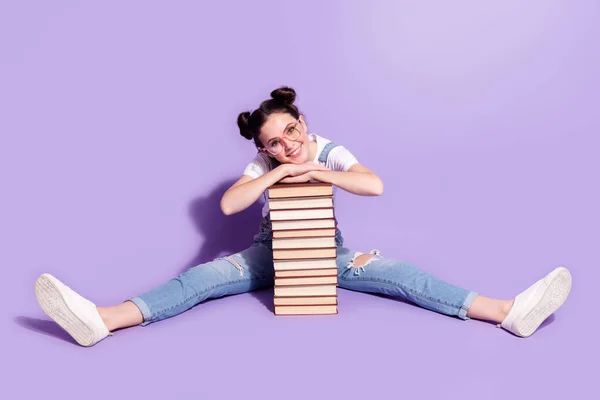 Foto von glänzenden niedlichen jungen Frau gekleidet Jeans Gesamtbrille sitzen Boden Arme Hände Bücher isoliert lila Farbe Hintergrund — Stockfoto