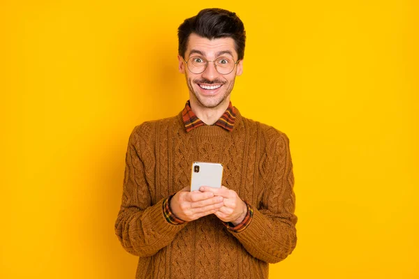 Heyecanlı adamın fotoğrafı, elinde telefon beyaz gülüşü, kameraya bak, gözlük tak, kahverengi kazak, parlak sarı arka plan. — Stok fotoğraf
