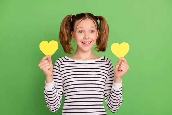 Foto de engraçado muito pequena estudante vestida camisola listrada segurando dois corações amarelos isolado fundo cor verde — Fotografia de Stock
