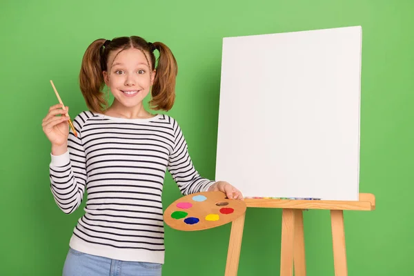 Zdjęcie imponujący ogon fryzura mała dziewczynka trzymać pędzel farba nosić sweter w paski izolowane na zielonym tle koloru — Zdjęcie stockowe