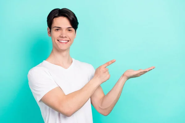 Foto av trevlig brunett frisyr tonåring kille hålla punkt tomt utrymme bära vit t-shirt isolerad på pulserande teal färg bakgrund — Stockfoto