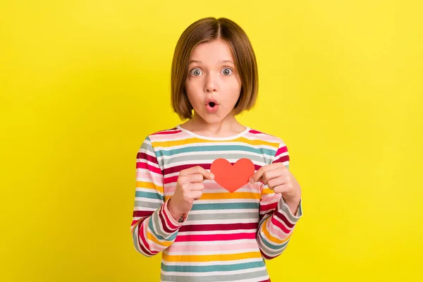 Foto de surpreendido chocado menina pequena segurar as mãos pequeno papel vermelho coração forma isolada no fundo de cor amarela — Fotografia de Stock
