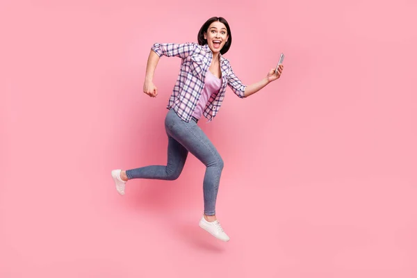 Photo de jeune femme funky brillant habillé chemise à carreaux souriant saut en cours d'exécution tapant gadget moderne isolé fond de couleur rose — Photo