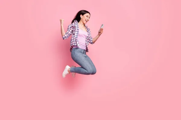 Pleine longueur photo de brillante jeune femme chanceuse porter chemise à carreaux saut à la recherche dispositif moderne levant poing isolé fond de couleur rose — Photo