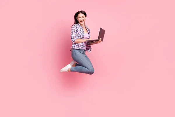 Foto de comprimento total de doce linda senhora vestida camisa quadriculada segurando gadget moderno saltando alto isolado cor de fundo rosa — Fotografia de Stock