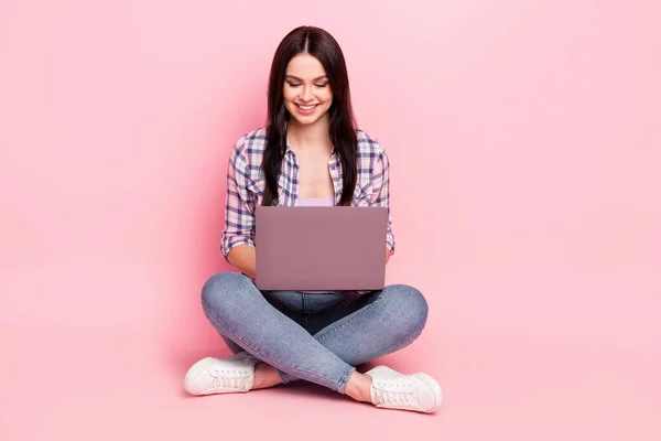 Full length body size photo girl in checkered shirt smiling sitting working on laptop odizolowany pastelowy różowy kolor tło — Zdjęcie stockowe