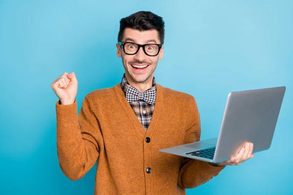 Foto de bom humor sorte jovem vestido casaco de lã marrom segurando dispositivo moderno levantando punho isolado azul cor de fundo — Fotografia de Stock