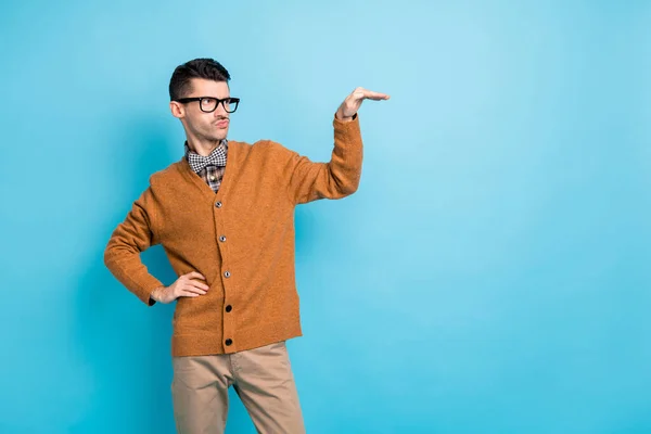 Foto de jovem engraçado duvidoso vestido marrom cardigan óculos medida braço vazio espaço isolado cor azul fundo — Fotografia de Stock