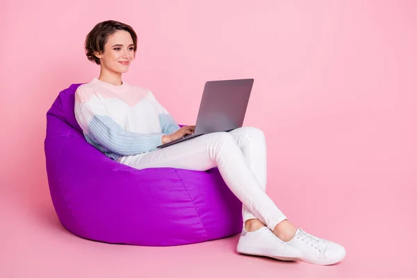 Potret wanita yang bekerja di atas laptop yang duduk di kursi beanbag ungu terisolasi di latar belakang berwarna merah muda pastel — Stok Foto