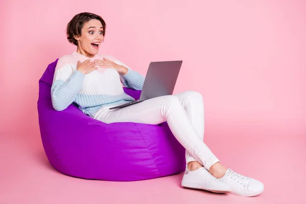 Potret panjang penuh dari wanita yang kagum dengan laptop yang duduk di kursi beanbag ungu terisolasi di latar belakang berwarna merah muda pastel — Stok Foto