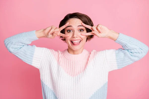 Retrato fotográfico de una chica excitada mostrando la lengua haciendo dos signos en V cerca de los ojos aislados sobre un fondo de color rosa pastel —  Fotos de Stock