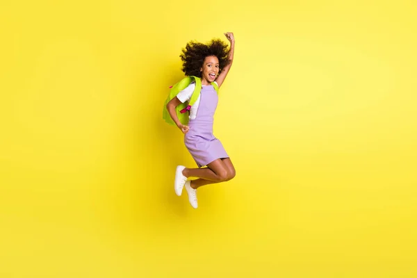 Фотография забавной школьницы в полный рост, прыгающей высоко, как победитель, в рюкзаке на ярко-желтом фоне — стоковое фото