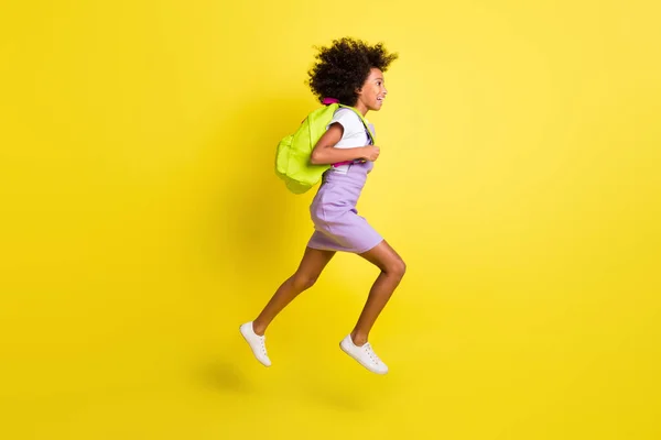 Full length body size profile side view of cheerful wavy-haired girl jumping running to lesson isolated over bright yellow color background