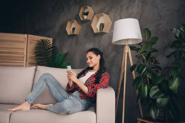 Retrato de chica atractiva alegre centrado sentado en el sofá descalzo utilizando gadget charlando en el loft moderno casa plana industrial en el interior — Foto de Stock