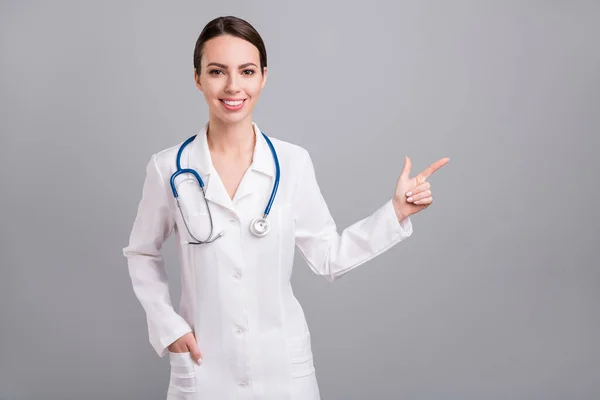 Fénykép fényes csinos fiatal hölgy orvos munkaruha mutató ujj üres tér mosolygós elszigetelt szürke színű háttér — Stock Fotó