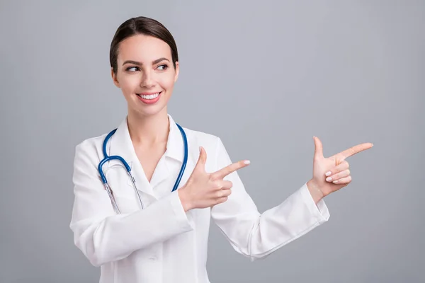 Photo of pretty charming young lady doctor workwear looking pointing fingers empty space smiling isolated grey color background — Stock Photo, Image