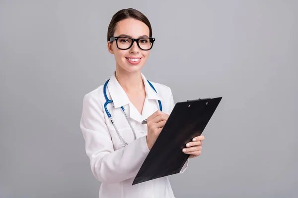 Foto von niedlich fröhlich junge doc Frau gekleidet weißen Mantel Brille Schreiben Klemmbrett lächeln isoliert graue Farbe Hintergrund — Stockfoto