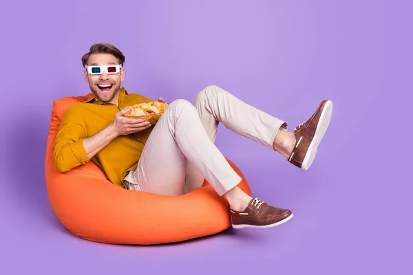 Foto di bello impressionato gentiluomo vestito camicia seduta beanbag guardare film mangiare patatine isolato colore viola sfondo — Foto Stock