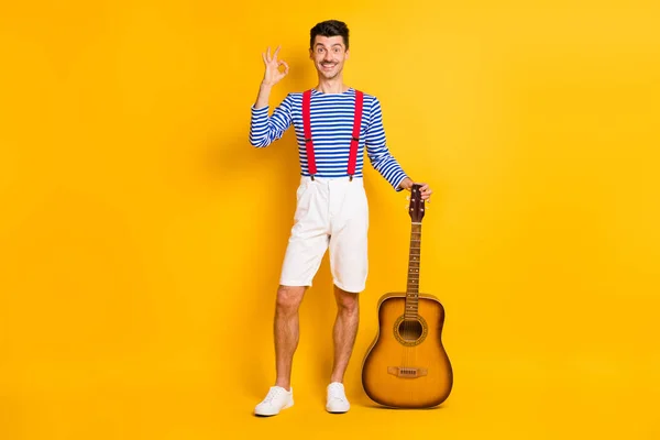 Full size zdjęcie młody przystojny wesoły dobry nastrój szczęśliwy człowiek pokaż okay znak trzymać gitarę izolowane na żółtym tle kolor — Zdjęcie stockowe